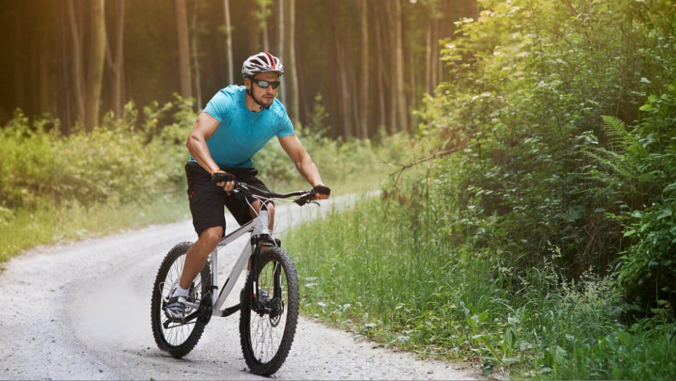Mężczyzna w niebieskiej koszulce, kasku i okularach przeciwsłonecznych jedzie na rowerze górskim po krętej, szutrowej ścieżce przez słoneczny las.